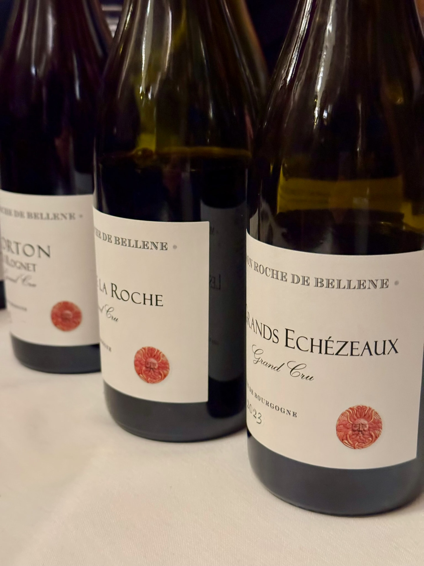Three Grand Cru Burgundy bottles from Maison Roche de Bellene arranged on a white tablecloth at a dimly lit tasting.