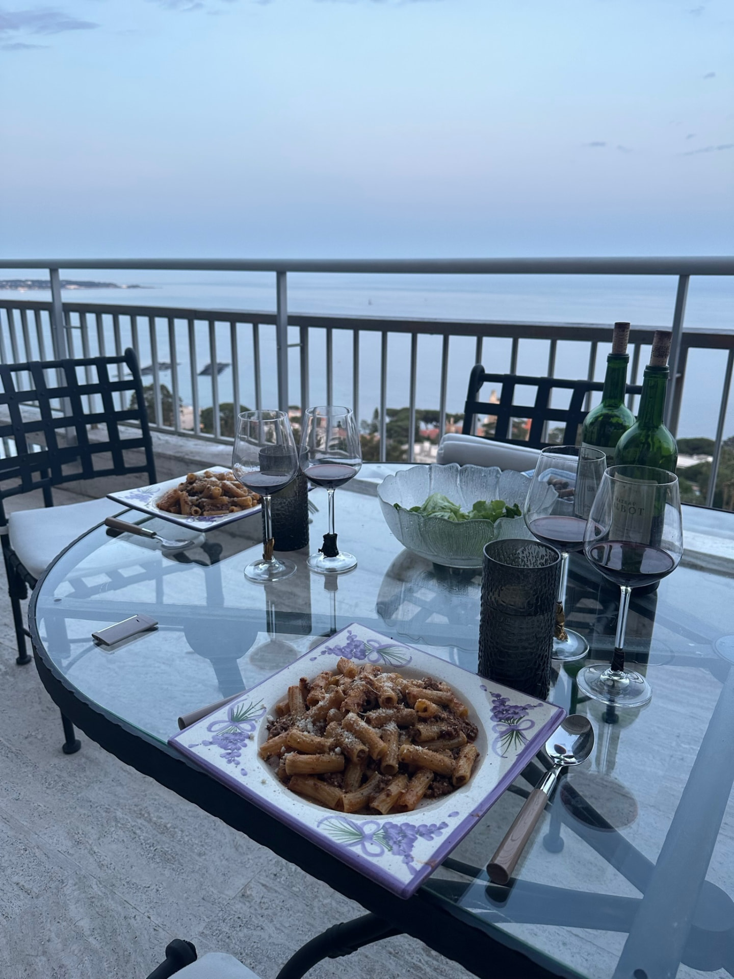 Dinner on a balcony overlooking the sea in Cannes: plates of pasta, glasses of red wine and two empty Bordeaux bottles at dusk.